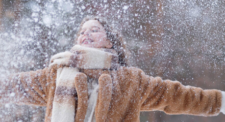 A white blonde girl in a knitted hat, a long scarf and a fur coat walks through the forest and smiles. Woman catches snow in white mittens. Winter is on the street. High quality photo. Christmas Eve