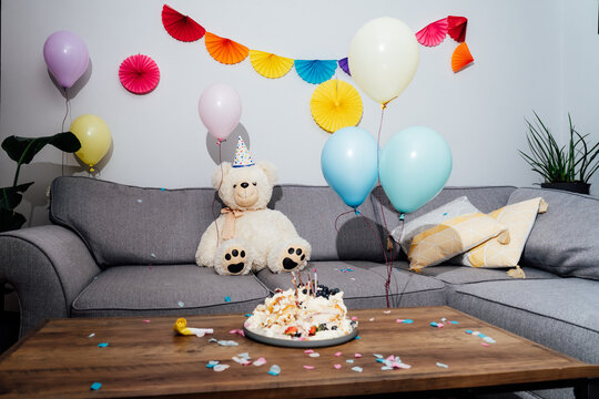 Messy table in living room after party. Broken birthday cake, confetti, balloons and decoration. Big teddy bear sitting on the sofa. Selective focus.