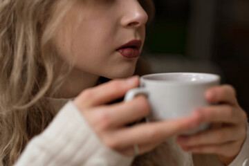 White blonde girl in a warm sweater drinks tea in a coffee shop. In a cafe, coffee is poured into a white mug. Hot tea for morning breakfast. Warming coffee in the restaurant on a winter evening. High