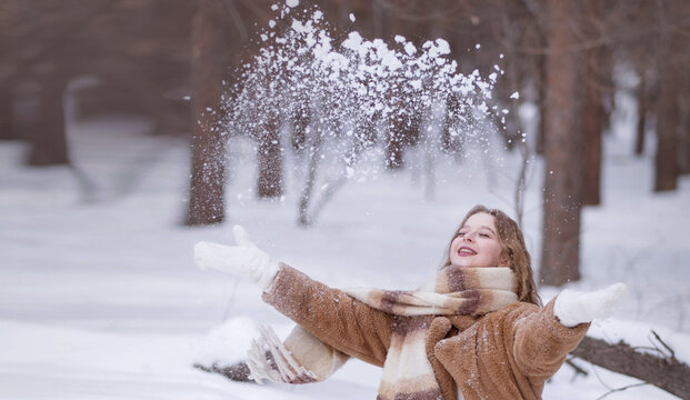 A white blonde girl in a knitted hat, a long scarf and a fur coat walks through the forest and smiles. Woman catches snow in white mittens. Winter is on the street. High quality photo. Christmas Eve