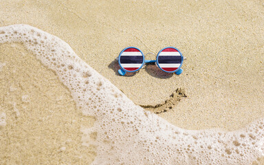Fototapeta na wymiar Sunglasses with flag of Thailand on a sandy beach. Nearby is a sea lightning and a painted smile. The concept of a successful vacation in the resorts of the Thailand.