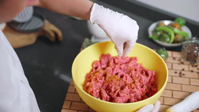 Woman in gloves seasoning raw mixed mince with aromatic spices powder in yellow bowl. Salt, pepper and paprika powder adding before making burgers, cutlets or meatballs. Aromatic ingredients for meat