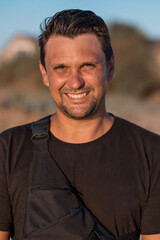 A handsome brunette man in a black t-shirt smiles and looks at the camera against the backdrop of mountains and a blue sky. High quality photo.