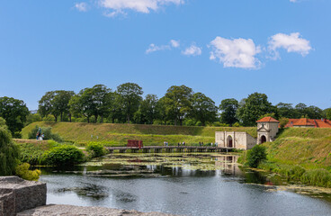 Fototapeta na wymiar Copenhagen, Denmark - July 24, 2022: Landscape, Green dirt ramparts and moat east of south entrance with bridge into historic military base Kastellet under blue sky. Red roofs