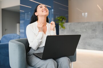 Portrait of excited young caucasian woman celebrating success while sitting with laptop at home or...