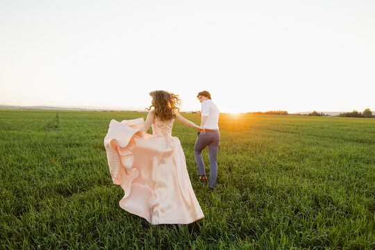 Teenage couple in prom attire running in rural field at sunset