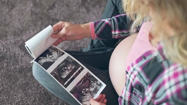 Expecting mother stroking her beautiful pregnant belly and looking on ultrasound scan pictures of her baby child, medical examination of baby gender reveal in prenatal period of pregnancy, happiness