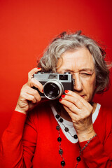 Portrait of a senior woman wearing red clothes, taking pictures with an old film camera, over a red background