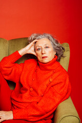 Portrait of a senior woman wearing red clothes, sitting on a green armchair, over a red background
