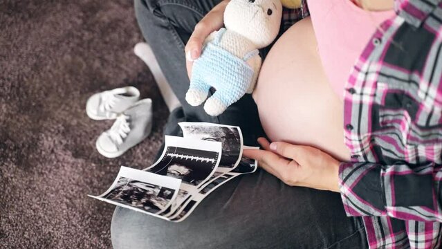 Happy pregnancy period of young woman, future mom sitting on the floor in room, holding baby scan pictures in hand and small knitted bunny toy, female with beautiful round belly waiting for her baby
