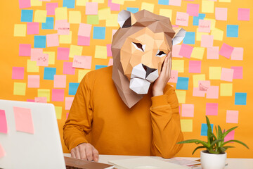 Horizontal shot of bored anonymous man in lion mask wearing orange sweater sitting at workplace with laptop against yellow wall with stickers, leaning on his head, tired from work.