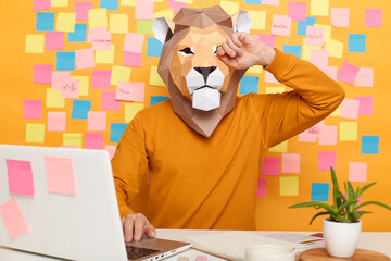 Image of exhausted anonymous man in lion mask wearing orange sweater sitting at workplace with...