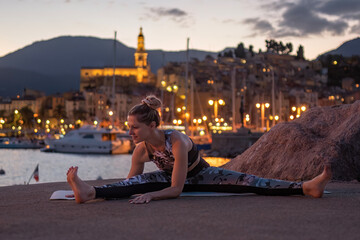 Fototapeta na wymiar full body portrait of a young woman doing a yoga workout on a stone feast during sunset and against the backdrop of a sea bay and blurred city with evening bokeh lights