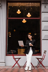 Cheerful girl with laptop standing near outside of cafe. Young woman is holding her laptop and smiling in cafe.