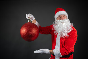 Portrait von einem verkleideten Weihnachtsmann mit riesiger Christbaumkugel in der Hand vor grauen...