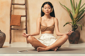 Asian woman in headphones meditating, sitting in yoga lotus pose, using meditation app on phone