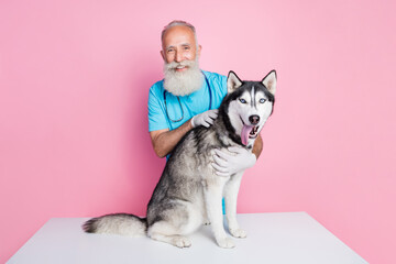 Photo of smiling positive elderly male vet wear blue uniform embracing big wolf dog isolated pink color background