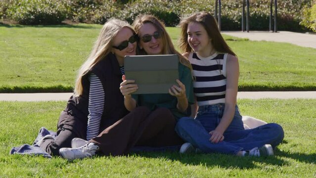Happy women enjoying sunny morning, sitting on green lawn. Cheerful female friends watching photos on a tablet. High quality 4k footage