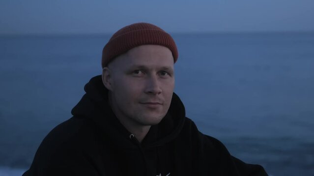 A man sits near the sea in the evening or at night and looks into the camera with a positive or kind look. Evening near the sea. High quality 4k footage