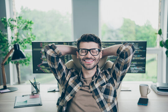 Photo of smiling positive programmer wear spectacles relaxing after working device hands behind head indoors workplace workshop home