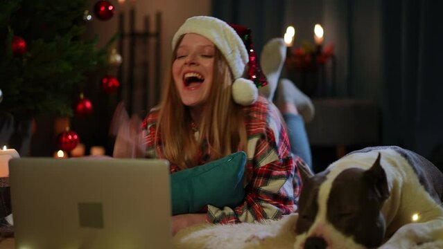Relaxed happy woman watching comedy movie online on laptop resting with dog in living room in darkness. Portrait of laughing Caucasian beautiful lady having fun on Christmas with pet indoors