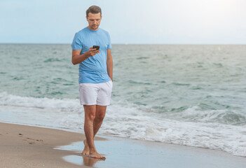 Young man holding mobile phone in his hand while walking on the beach