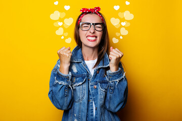 young woman in a hair band, in a denim jacket clenching her fists on a yellow background, closed her eyes.