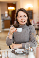 Dreamy brunette woman sitting at table in cozy empty cafe with cup of aromatic coffee during breakfast..