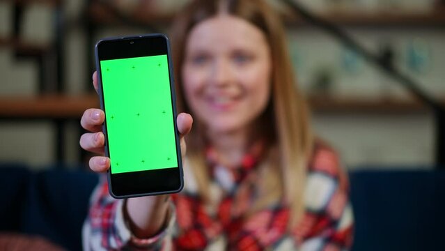 Close-up green screen smartphone in female Caucasian hand with blurred young woman smiling at background. Unrecognizable young lady showing website app template on phone indoors