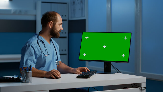 Medical assistant sitting at desk typing patient disease report on computer with green screen chroma key mock up display. Physician nurse working late at night in hospital office. Health care concept