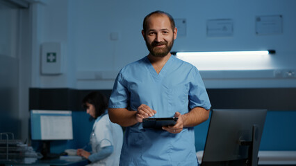 Physician nurse holding tablet computer checking patient diagnosis while typing medical expertise during night shift in hospital office. Assistant man in blue uniform working at health care treatment