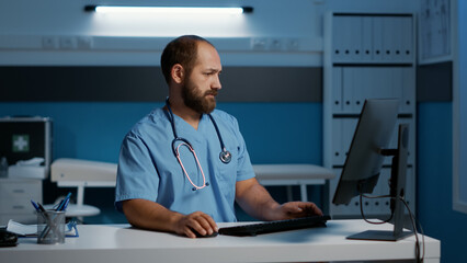 General practitioner nurse sitting at desk typing medical expertise on computer while working late at night at patient health care treatment in hospital office. Medicine service and concept