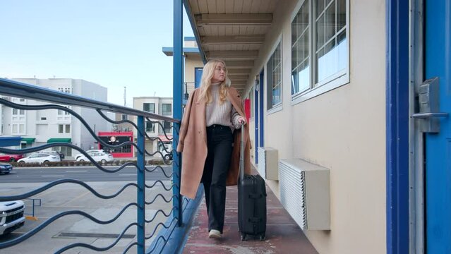 Caucasian, blonde girl strolling along motel corridor with a travel suitcase. Female traveler departing to the airport from a hotel complex. High quality 4k footage