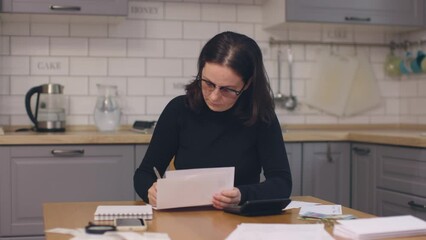 serious caucasian woman look through paper document letters signing them working kitchen table home indoors. female wear eyeglasses working at home paperwork reading papers writing down data - Powered by Adobe