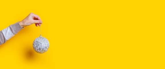 Female hand holds decorative Christmas ball on yellow background. Banner