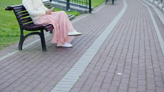 alone woman pink white clothes sit on wooden bench in city park using smartphone touching screen. female put mobile phone into pocket of white coat zip pocket stands up from bench. warm weather