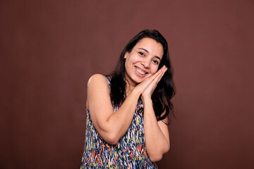 Smiling relaxed indian woman showing sleeping gesture, lying on folded hands, looking at camera. Cheerful positive model making sleepy sign, laughing, front view medium shot