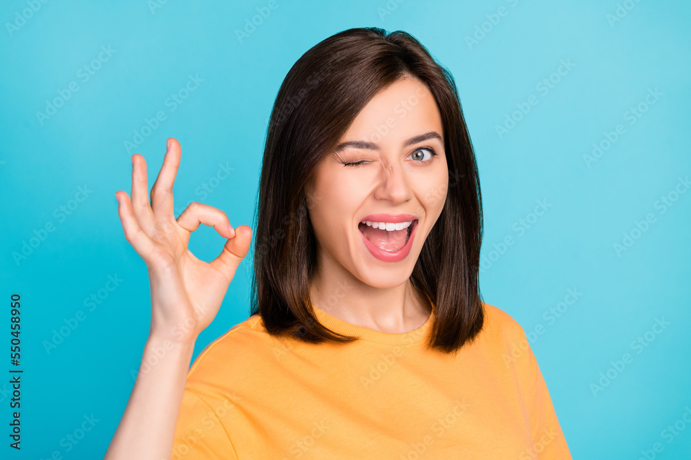 Sticker Photo of coquettish positive satisfied girl with straight hair wear yellow t-shirt showing okey winking isolated on blue color background