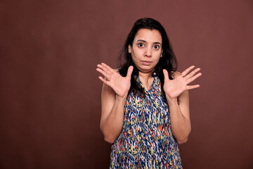 Indian woman showing no problem gesture with palms, looking at camera with confused facial expression. Young lady standing with spread arms portrait, front view studio medium shot on brown background
