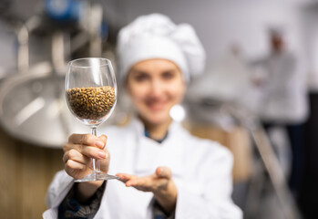 Skilled satisfied female brewer in white uniform demonstrating high quality malted barley grain in glass while standing at brewery, selective focus on glass with malt
