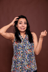 Fototapeta premium Thoughtful smiling indian woman rubbing head with confused facial expression portrait, awkward pose. Puzzled lady doubting, uncertain person thinking, looking at camera, studio medium shot
