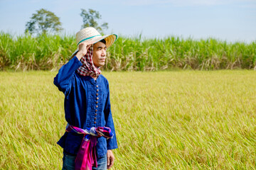 A Thai male farmer wearing a traditional blue shirt and hat is standing and posing holding his hat in the middle of the field.