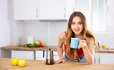 Portrait of relaxed young woman drinking coffee or tea in morning at kitchen