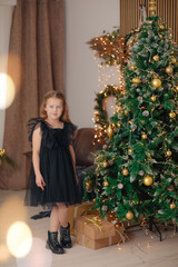 Beautiful little girl in a black dress near a fir tree. Christmas dress for girls. Fashionable girl is waiting for gifts under Christmas tree with bright lights