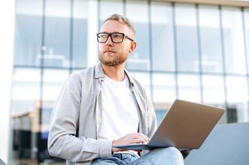 Serious focused caucasian bearded man with glasses, in casual clothes, sits outdoors near the office with a laptop, works on a project, looks away thoughtfully, plans a strategy, thinking over idea