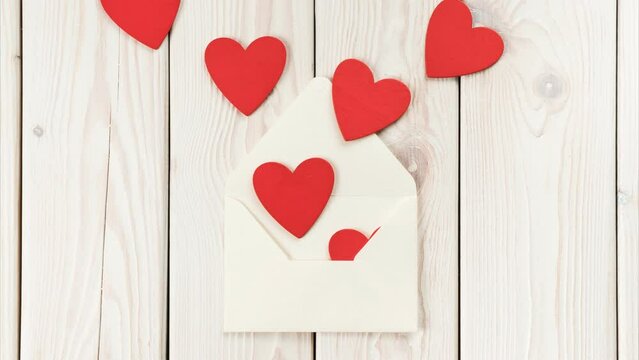 White paper craft envelope moving to the center and giving rain of red paper hearts on light wooden background. St Valentines, women or wedding celebration message, letter. Stop motion, copy space