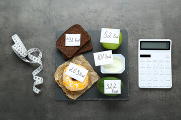 Calculator and food products with calorific value tags on dark grey table, flat lay. Weight loss...