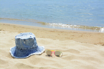 Fototapeta na wymiar Jeans hat and sunglasses on sand near sea, space for text. Beach accessories