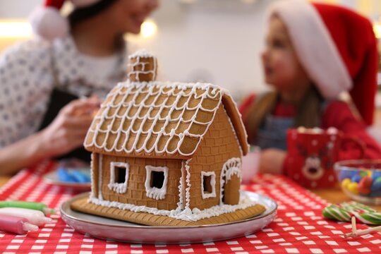 Mother and daughter in Santa hats cooking together, focus on gingerbread house