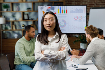 Confident happy smiling diverse asian woman, startup team leader, standing against background of mixed race office workers or business people with arms crossed or clasped, looking at camera.